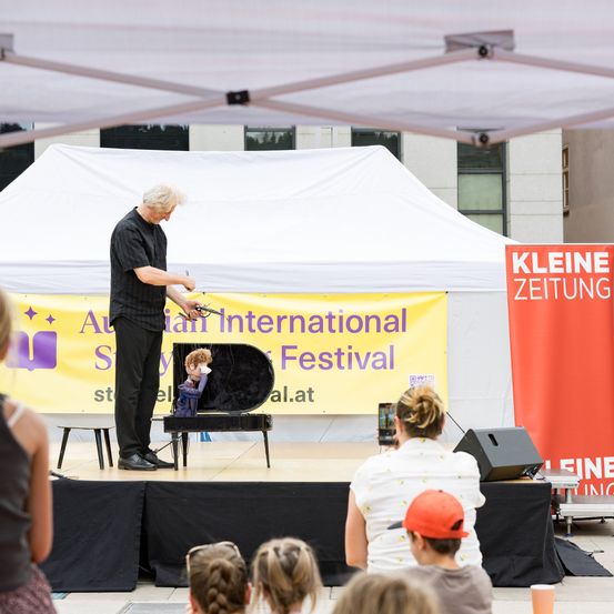 Ein Mann führt eine Puppenshow auf der Bühne beim Australian International Story Festival auf. Das Publikum beobachtet aus verschiedenen Positionen, und ein Banner hinter dem Darsteller trägt die Aufschrift 'Australian International Story Festival.'