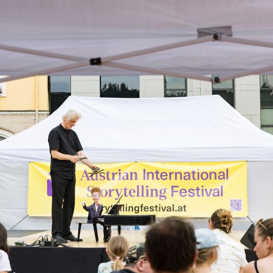 Ein Mann auf der Bühne beim Austrian International Storytelling Festival, der mit einer Marionette auftritt. Hinter ihm zeigt ein gelbes Banner den Namen der Veranstaltung. Das Publikum sitzt davor.