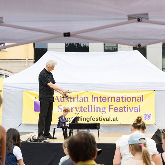 Ein älterer Mann führt eine Zauberkunst mit einer Puppe auf der Bühne unter einem Zelt vor, während ein Junge das Klavier spielt. Das Publikum beobachtet von vorne.