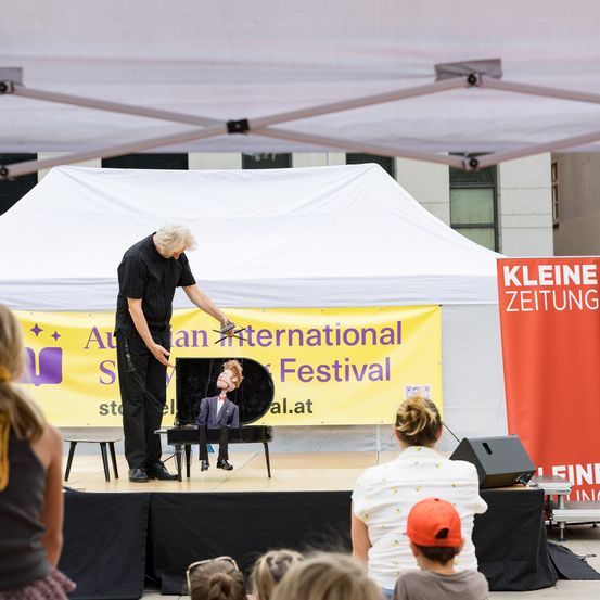 Ein Magier führt auf der Bühne eine Vorstellung mit einer Puppe in einem Klavier vor, unter einem Zelt, mit zuschauenden Zuschauern. Ein Banner dahinter lautet 'Australian International Festival'.