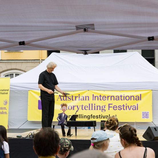 Ein Mann führt ein Puppenspiel auf der Bühne beim Austrian International Storytelling Festival auf. Er sitzt am Klavier, während eine Puppe auf der Bühne vor einem Publikum auftritt.