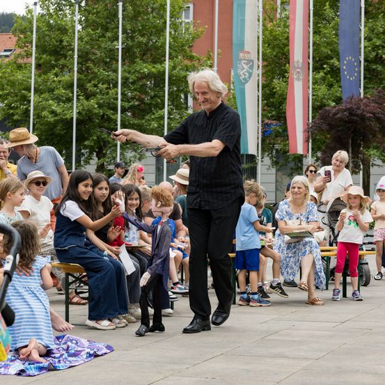 Ein älterer Mann unterhält eine Gruppe von Kindern mit einer Puppenshow in einem Park. Die Kinder sitzen auf Bänken, während andere stehen oder auf dem Boden sitzen. Dahinter sind Flaggen zu sehen.