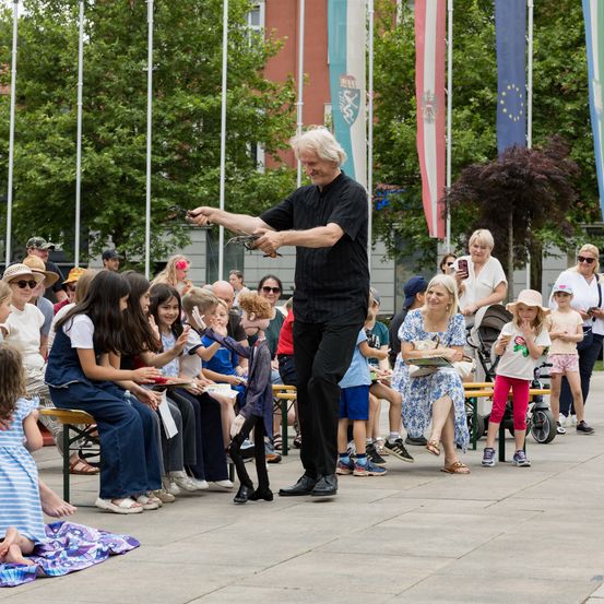 Ein Mann führt ein Puppenspiel für eine Menschenmenge aus Kindern und Erwachsenen in einem Park auf. Das Publikum sitzt auf Bänken, während der Mann vor ihnen steht.