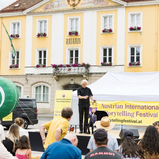 Ein älterer Mann erzählt Geschichten unter einem Zelt beim Austrian International Storytelling Festival vor einem Gebäude mit Blumen und einem Balkon.