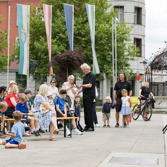 Mehrere Erwachsene und Kinder sitzen unter Flaggen auf Bänken in einem Stadtplatz, während zwei Männer mit ihnen interagieren. In der Nähe steht ein Fahrrad auf dem Boden.