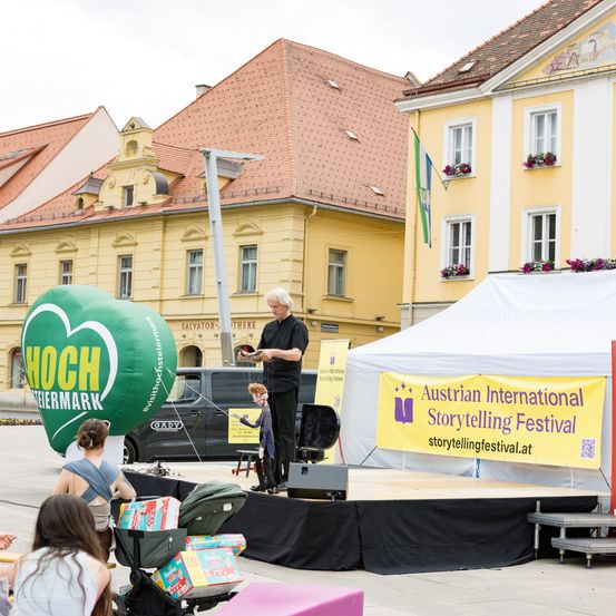 Ein Geschichtenerzähler steht auf der Bühne mit einem grünen Luftballon. Ein Banner steht für das Österreichische Internationale Erzählfestival. Gebäude mit roten Dächern und gelben Fassaden dahinter.