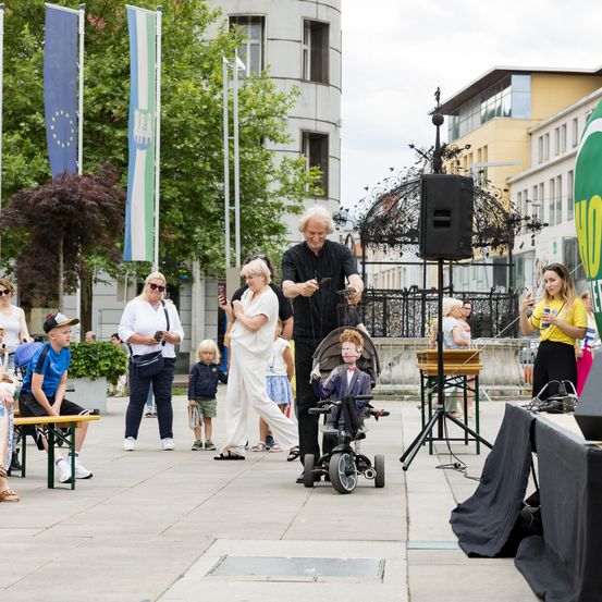 Ein Mann führt ein Puppenspiel mit einer Marionette im Anzug auf einem Dreirad vor einer Menge auf.