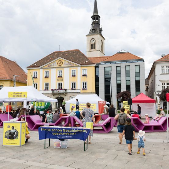 Ein Platz mit bunten Bänken und Zelten, mit einem Turm und Gebäuden im Hintergrund. Menschen gehen herum und sitzen auf den Bänken.