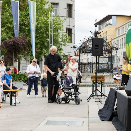 Ein Mann schiebt einen Kinderwagen, während ein anderer Mann filmt. Zuschauer, einschließlich einer Frau, die ein Foto macht, beobachten. Eine Bühne mit einem Lautsprecher ist im Hintergrund.
