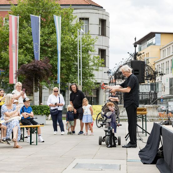 Ein Mann führt eine Zaubervorführung mit einer Spielzeugfigur vor einer Menge durch, einschließlich Kindern, auf einem Stadtplatz. Hinter ihm steht ein schwarzer Lautsprecher auf der Bühne.