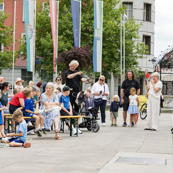 Eine Gruppe von Menschen, einschließlich Familien, sitzt auf Bänken unter Bäumen, einige auf einem gepflasterten Gehweg. Ein Mann steht und führt auf der Bühne mit Flaggen im Hintergrund auf.