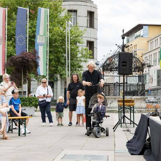 Ein Mann führt auf der Straße eine Puppenshow vor, während eine Menschenmenge zusieht. Er hat eine Puppe in den Händen. Es gibt einen Lautsprecher auf einem Stativ, und im Hintergrund sind einige Fahnen zu sehen.