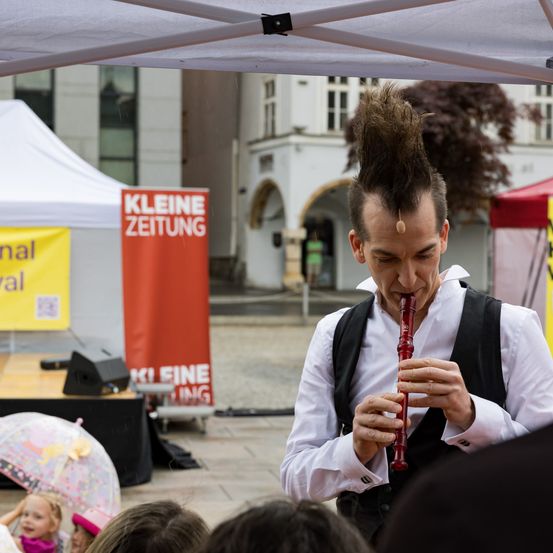 Ein Mann mit spitzem Haar spielt draußen unter einem Zelt eine rote Blockflöte. Ein Schild hinter ihm sagt Kleine Zeitung. Leute beobachten ihn, darunter ein Mädchen mit einem Regenschirm.