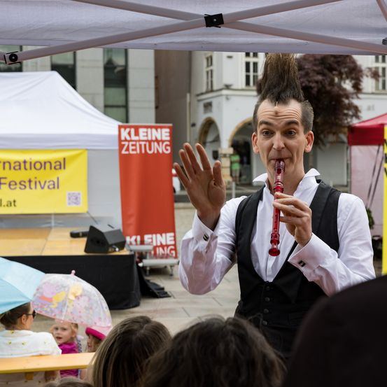 Ein Künstler spielt eine rote Blockflöte auf der Bühne bei einem Festival, vor einem Publikum. Banner für das 'Internationale Festival' und 'Kleine Zeitung' sind sichtbar.
