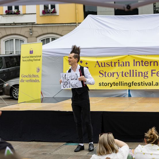 Ein Mann steht auf der Bühne mit einem Schild, das 'HELLO BRICK' steht. Ein Zelt steht hinter ihm mit den Worten 'Austrian International Storytelling Festival'. Ein Banner an der Seite sagt 'Geschichten, sie verbinden'. Eine Frau beobachtet in der Nähe.