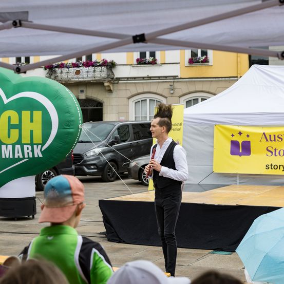 Ein Straßenkünstler spielt eine Flöte auf einer Bühne, mit einem Ballon und einem Zelt dahinter. Ein Publikum schaut zu, darunter ein Kind mit einer rosa Mütze.