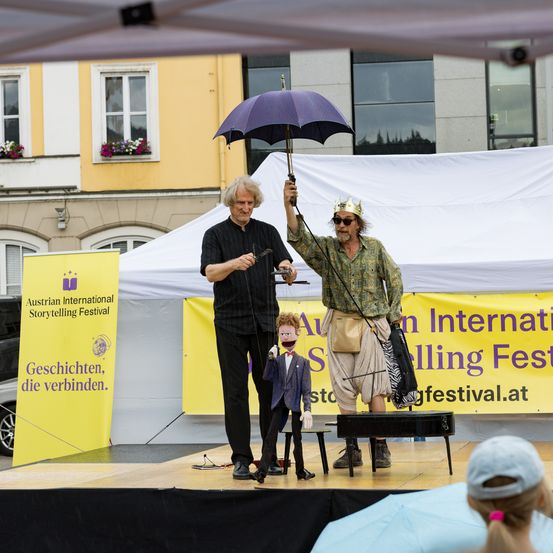 Zwei Männer führen mit einer Marionette beim Österreichischen Internationalen Storytelling-Festival auf. Einer hält einen Schirm und der andere eine Marionette mit Krone.