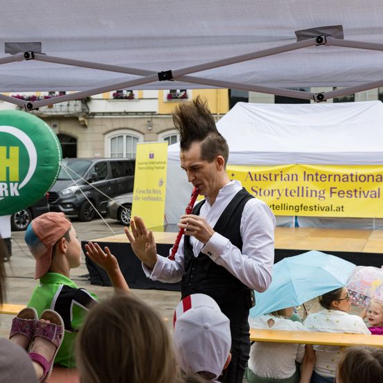 Ein Mann mit spitzem Haar spielt eine rote Blockflöte beim Austrian International Storytelling Festival unter einem Zelt. Ein Kind und Erwachsene lauschen ihm.