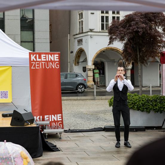 Ein Mann steht auf einer Bühne mit einem Schild, das Kleine Zeitung sagt. Er spielt ein Instrument. Ein Auto ist auf der Straße geparkt.