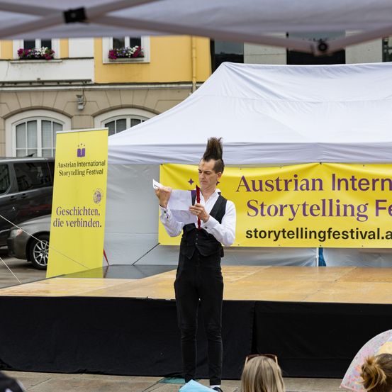 Ein Mann steht auf einer Bühne und hält ein Papier in der Hand, unter einem Zelt mit einem Banner, das das Internationale Österreichische Erzählfestival bewirbt.