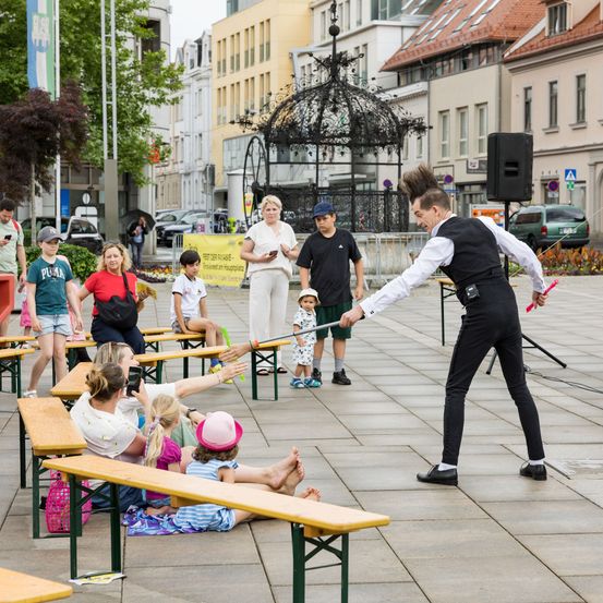 Ein Mann in einem schwarzen Anzug führt eine Schwertschlucker-Vorstellung auf einem Stadtplatz durch, während Menschen auf Bänken sitzen und zusehen. Gebäude und ein Pavillon sind im Hintergrund.