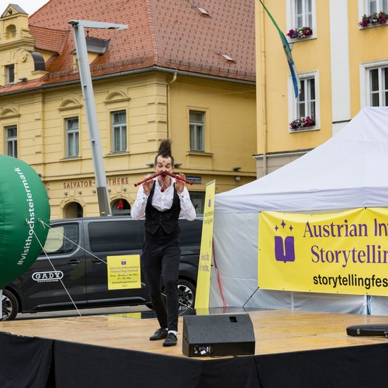 Ein Mann tritt auf einer Bühne mit einem Mikrofon auf, trägt einen schwarzen Anzug. Hinter ihm steht ein Zelt und ein gelbes Banner, das für das Austrian International Storytelling Festival wirbt. Im Hintergrund befinden sich Gebäude und ein grüner Ballon.