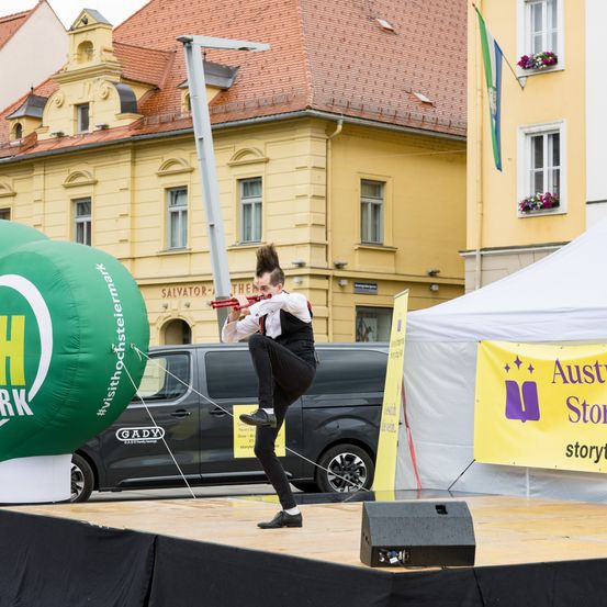 Ein Künstler steht auf der Bühne und hält eine Trompete, vor einem gelben Banner. Ein grüner Ballon mit Text und ein großes gelbes Gebäude stehen hinter dem Künstler.