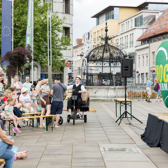Ein Mann tritt auf einem Einrad auf einem Stadtplatz auf. Zuschauer schauen zu, einige auf Bänken und andere stehend. Die Umgebung umfasst Bäume, Flaggen und eine Laube.