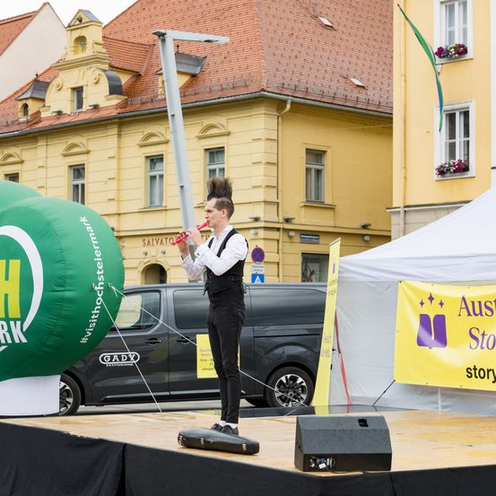 Ein Mann in weißem Hemd und schwarzer Hose spielt auf der Bühne eine Flöte mit einem großen grünen Luftballon im Hintergrund. Ein Van ist hinter ihm geparkt und ein Zelt steht rechts.