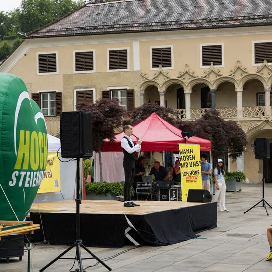 Ein Mann steht auf einer Bühne mit einem Mikrofon auf einem öffentlichen Platz. Hinter ihm steht ein rotes Zelt, und es gibt ein gelbes Banner mit Text. Ein aufblasbarer grüner Ballon mit Text ist links.
