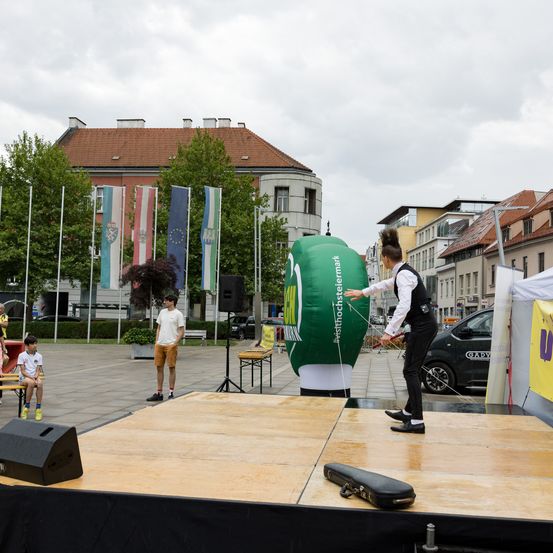 Ein Mann führt eine Zaubervorführung auf einer Bühne vor einem Publikum durch. Ein riesiger grüner Ballon ist anwesend und Flaggen im Hintergrund.