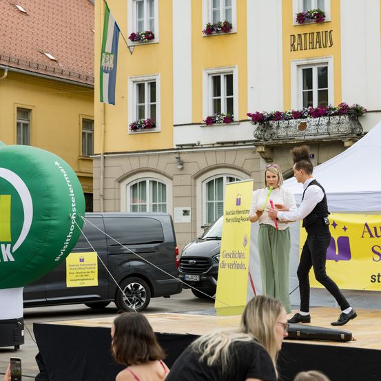 Eine Bühne mit einem grünen Luftballon und zwei Künstlern vor einem gelben Gebäude mit Balkon und Flagge, wo ein Publikum zuschaut.