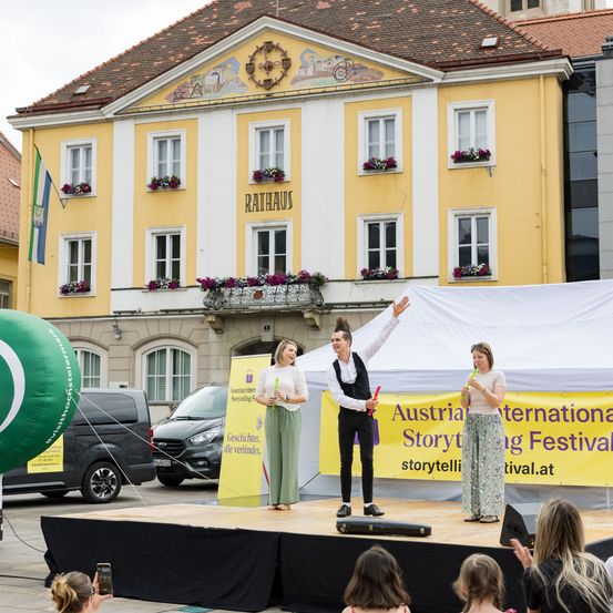 Eine Bühne mit Darstellern vor einem gelben Gebäude mit der Aufschrift 'Rathaus', ein Publikum schaut zu, und ein grüner Luftballon mit einer Flagge ist vorhanden.