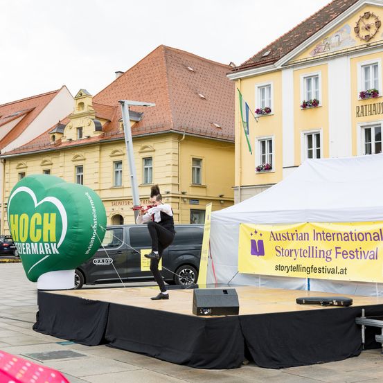 Eine Person tritt auf einer Bühne vor einem herzförmigen Luftballon und einem gelben Banner auf, das für das Österreichische Internationale Erzählkunstfestival wirbt. Gebäude mit roten Dächern und Fahnen sind im Hintergrund.