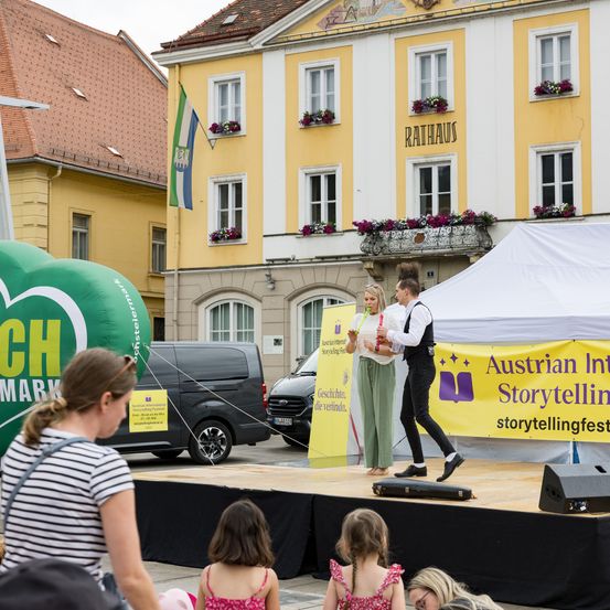 Eine Bühne mit einem gelben Banner, das 'Austrian International Storytelling' liest, mit zwei Darstellern. Ein Publikum schaut zu, und ein Van ist in der Nähe geparkt.