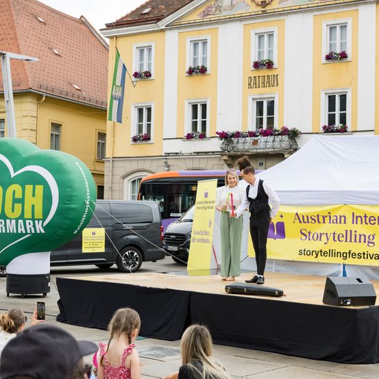 Ein Storytelling-Event findet auf dem Stadtplatz statt. Eine Frau und ein Mann stehen auf der Bühne, halten Bücher in den Händen, während eine Menge zusieht. Dahinter sind ein Bus und ein Lieferwagen geparkt, und ein großer Ballon befindet sich auf der linken Seite.