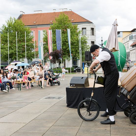 Ein Mann fährt mit einem Einrad und einem Anhänger voller Gepäck in einer Stadtplatz, wobei eine Menschenmenge zusieht.