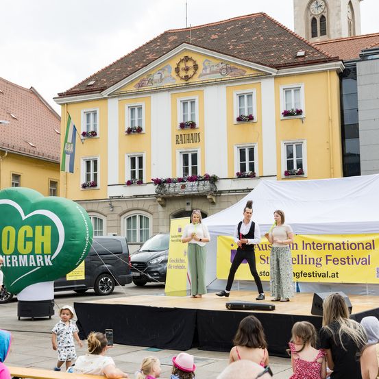 Drei Personen treten vor einem Publikum auf der Bühne auf. Ein gelbes Banner mit dem Text 'Austen International Storytelling Festival' befindet sich dahinter. Ein grüner Ballon in Herzform steht links. Ein Gebäude mit mehreren Fenstern befindet sich hinter der Bühne. Autos sind vor dem Gebäude geparkt.
