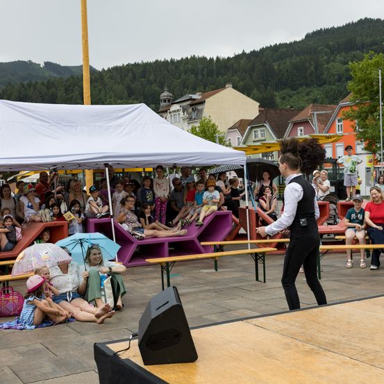 Ein Künstler auf der Bühne unterhält eine Menschenmenge aus Erwachsenen und Kindern im Freien. Ein Zelt steht hinter dem Publikum.
