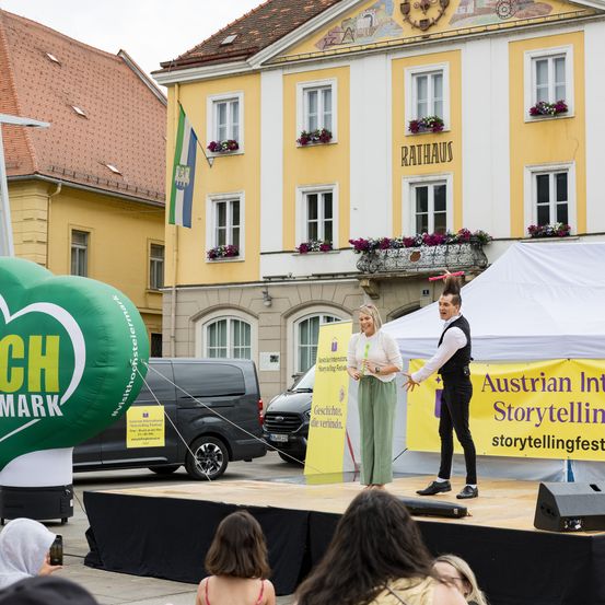 Zwei Personen stehen auf einer Bühne vor einem gelben Gebäude mit dem Schild Rathaus. Ein grüner Luftballon mit dem Wort 'CH' ist neben ihnen. Eine Menge beobachtet sie.