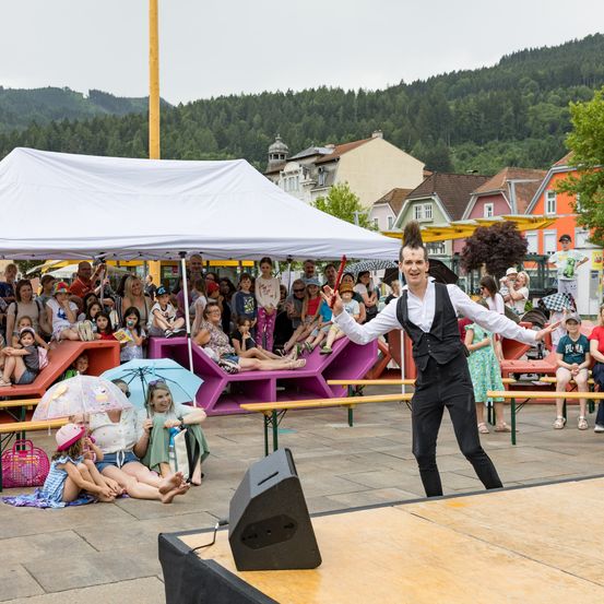 Ein Mann tritt auf der Bühne mit einem Mikrofon auf, während eine Menge zuschaut. Die Menge sitzt und steht unter Zelten. Einige Kinder sitzen auf dem Boden.