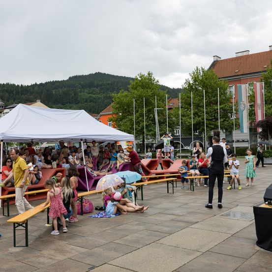 Unter einem weißen Zelt sitzen eine Gruppe von Kindern und Erwachsenen auf Bänken und beobachten eine Aufführung. In der Nähe steht ein Mann auf der Bühne. Im Hintergrund befinden sich Bäume, ein Gebäude und eine Bergkette.