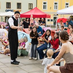 Ein Straßenkünstler unterhält eine Menge von Kindern und Erwachsenen. Einige sitzen auf Bänken, andere stehen unter Regenschirmen, alle schauen zu und klatschen.