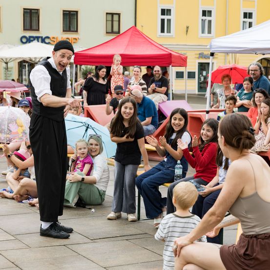 Ein Straßenkünstler unterhält eine Menge von Kindern und Erwachsenen. Einige sitzen auf Bänken, andere stehen unter Regenschirmen, alle schauen zu und klatschen.