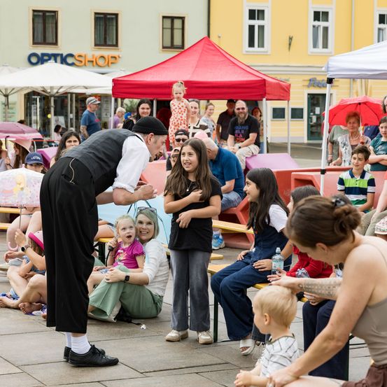 Ein Straßenkünstler unterhält eine Gruppe von Kindern und Erwachsenen im Freien. Zuschauer sitzen auf Bänken unter Zelten und beobachten die Vorstellung.