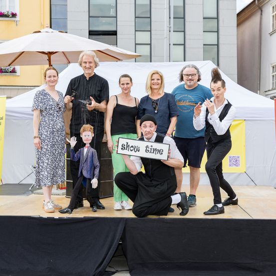 Eine Gruppe von Menschen, einschließlich Puppenspieler, stehen auf einer Bühne mit einem kleinen Puppen, die ein Schild mit der Aufschrift Show Time hält. Dahinter befindet sich ein großes weißes Zelt mit einem Gebäude im Hintergrund.