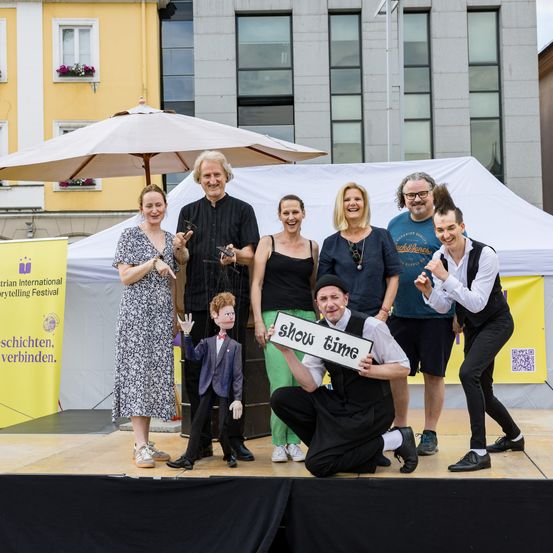 Gruppe von Menschen auf der Bühne bei einem Erzählfestival, einer hält ein Schild mit der Aufschrift 'show time' und einer Puppe. Dahinter ein gelber Banner mit fremdem Text und einem Gebäude mit Fenstern und Blumen.