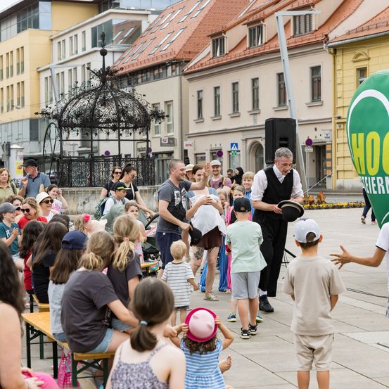 Eine Straßenaufführung zieht eine Menschenmenge in einem Stadtplatz an, einschließlich Kinder und Erwachsene. Ein Mann in einem Weste hält einen Hut. Gebäude und ein Pavillon sind im Hintergrund.