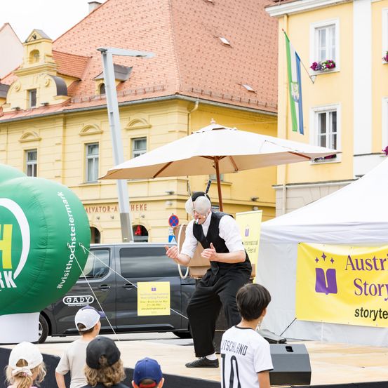 Ein Künstler unterhält eine Gruppe von Kindern auf der Bühne. Die Umgebung umfasst ein gelbes Zelt, ein gelbes Gebäude mit rotem Dach und einen Ballon.