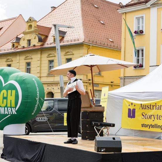Ein Mann steht auf der Bühne mit einem Schild und einem Schirm vor Gebäuden. Zwei grüne Luftballons sind auf der Bühne. Ein Zelt mit einem gelben Banner ist hinter ihm.